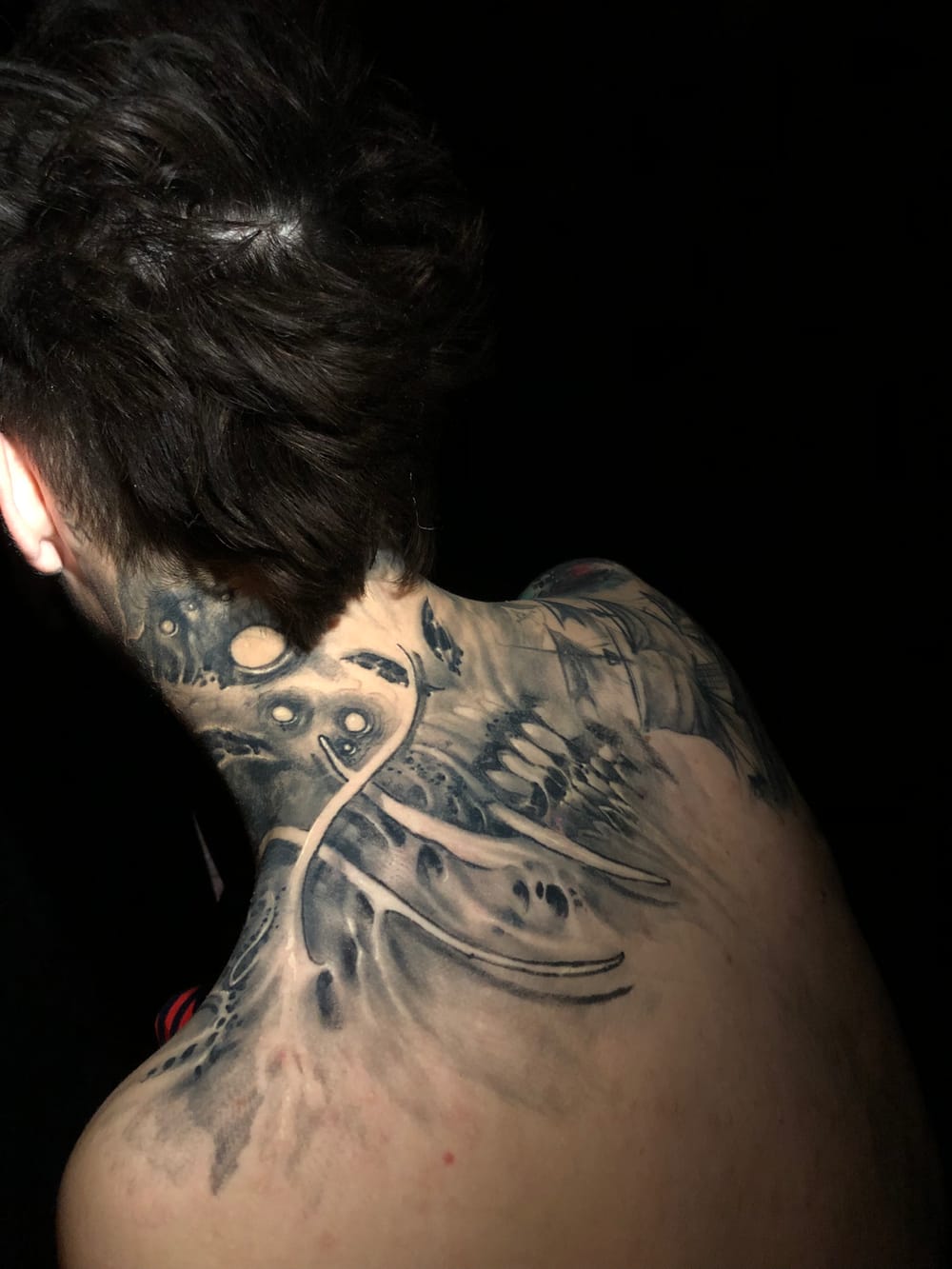 Dark surreal black & grey tattoo across the neck and back — spectral faces emerging from smoke