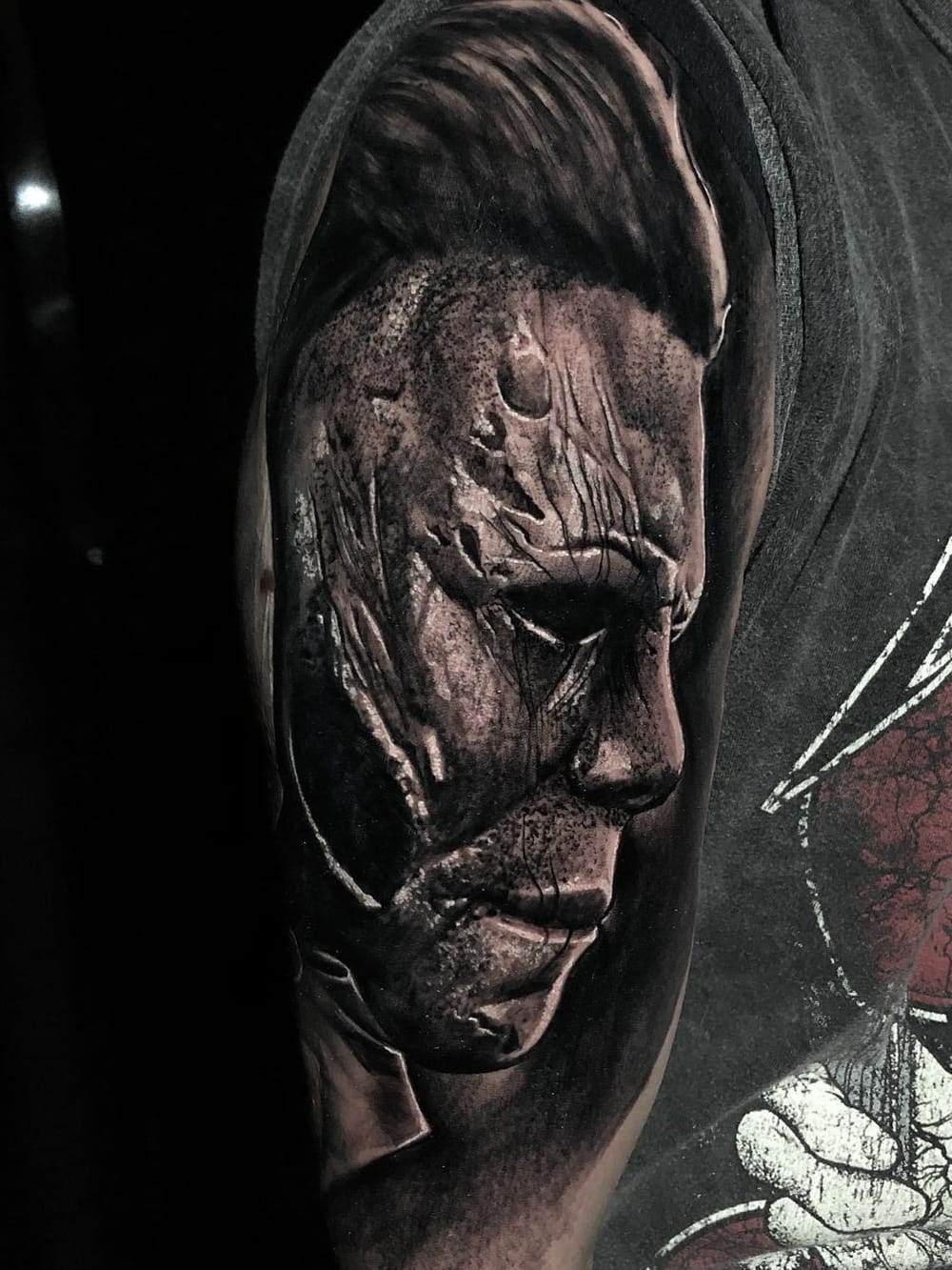 Black & grey horror realism tattoo — a cinematic masked figure portrait on the upper arm